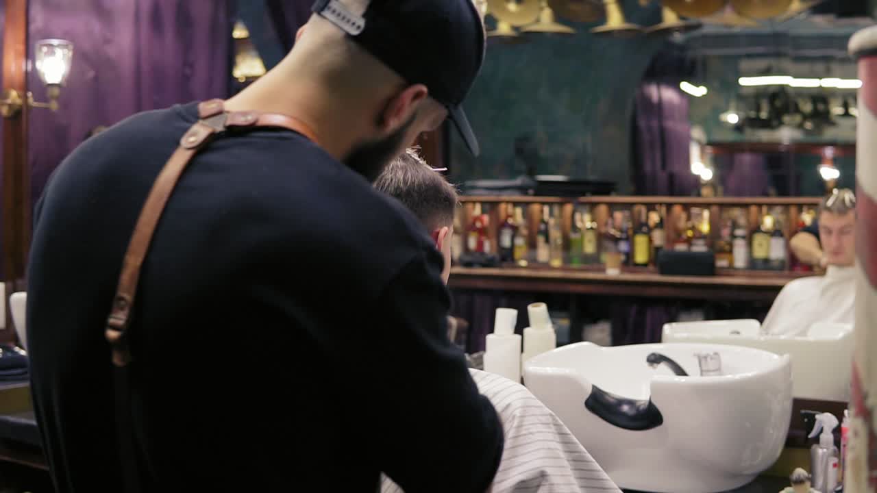 Back view of a bearded barber cutting hair using electric trimmer on the neck of his customer in barber shot. Slow Motion shot