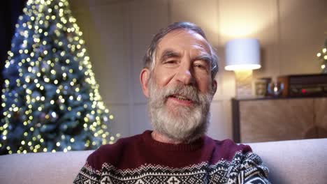 Close-Up-Of-Happy-Old-Male-Pensioner-In-Positive-Mood-Video-Chatting-Near-Glowing-Decorated-Xmas-Tree