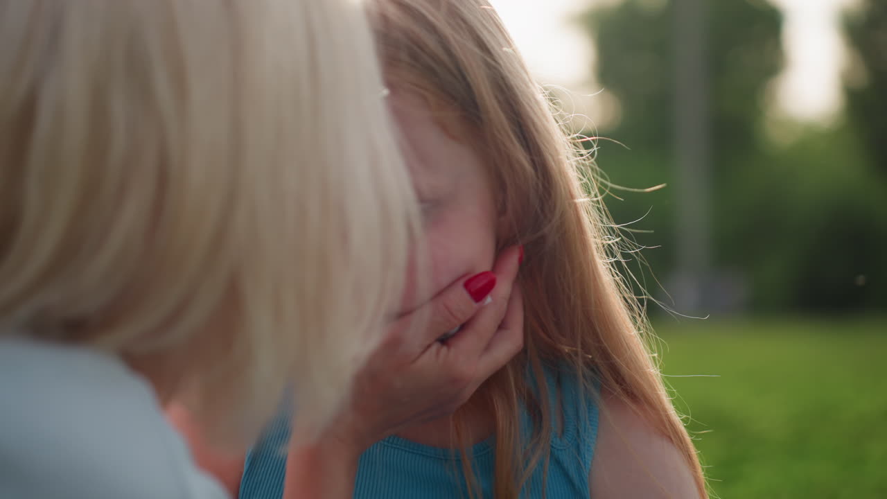 joyful mother with sunglasses cleaning daughter face with tissue seated on grass under warm sunset light, soft background blur highlighting loving interaction and playful family bonding outdoors