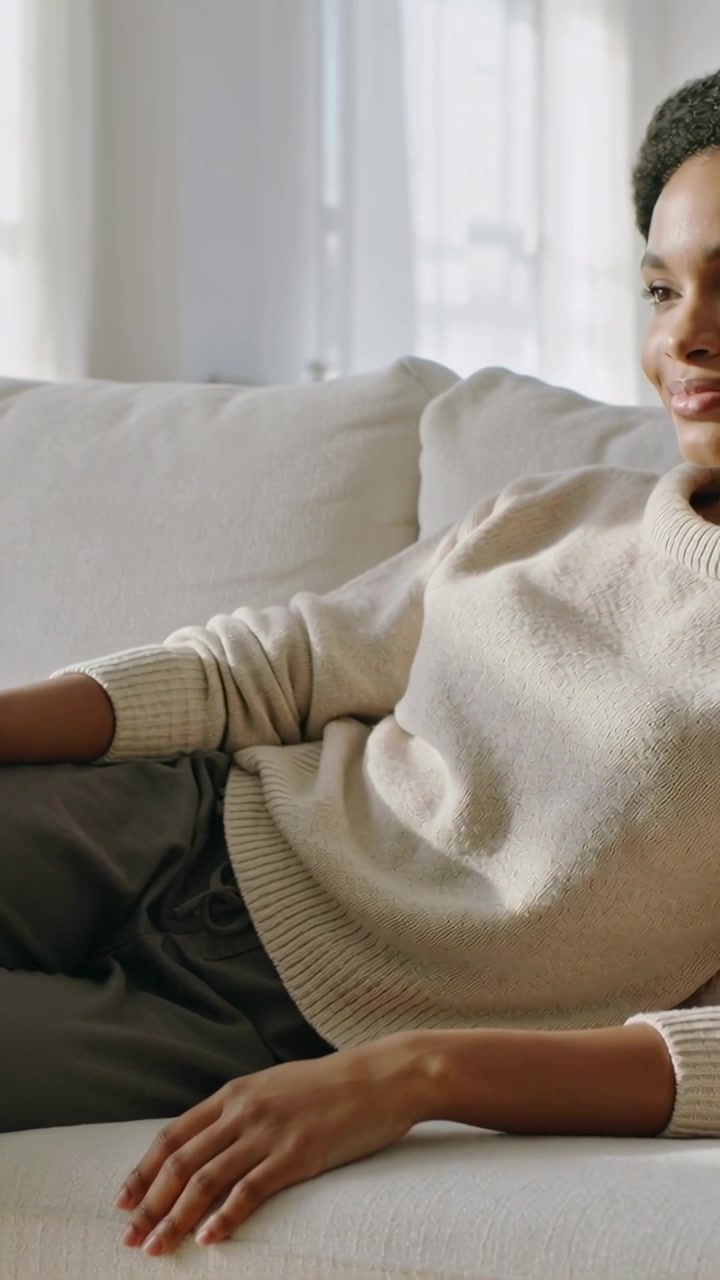 Vidéo verticale : Femme se relaxant sur un canapé en pull, s'installant tandis qu'une lumière douce filtre à travers les rideaux.