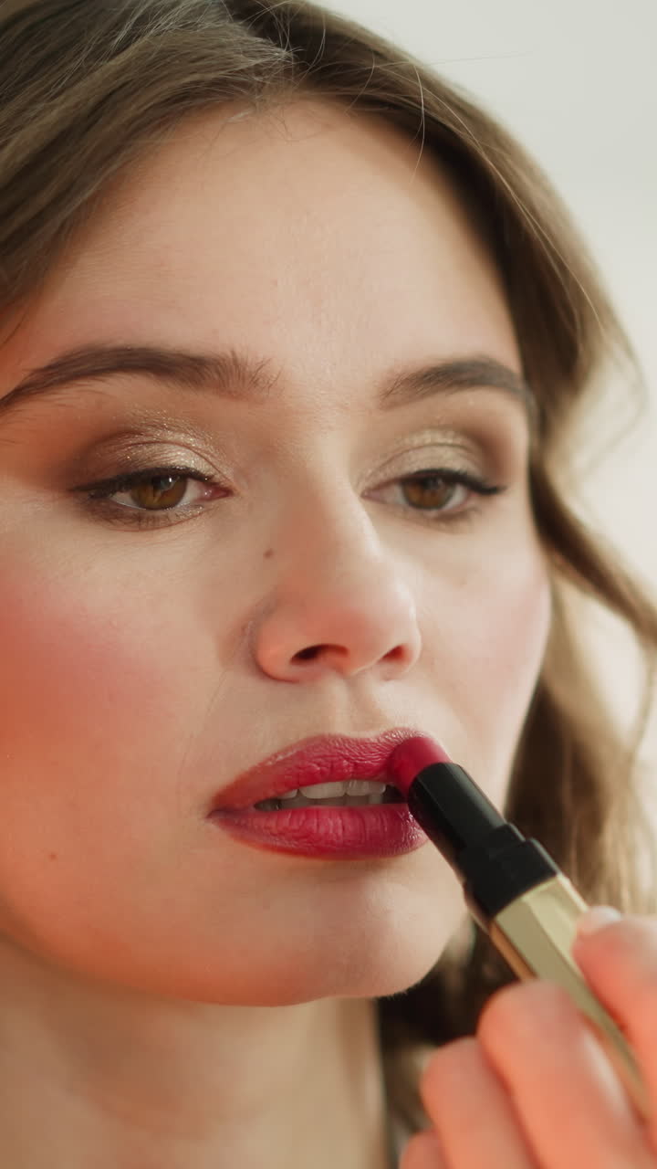 Focused young woman paints lips with glamorous red lipstick looking in mirror. Female person does bright makeup to go to birthday party closeup