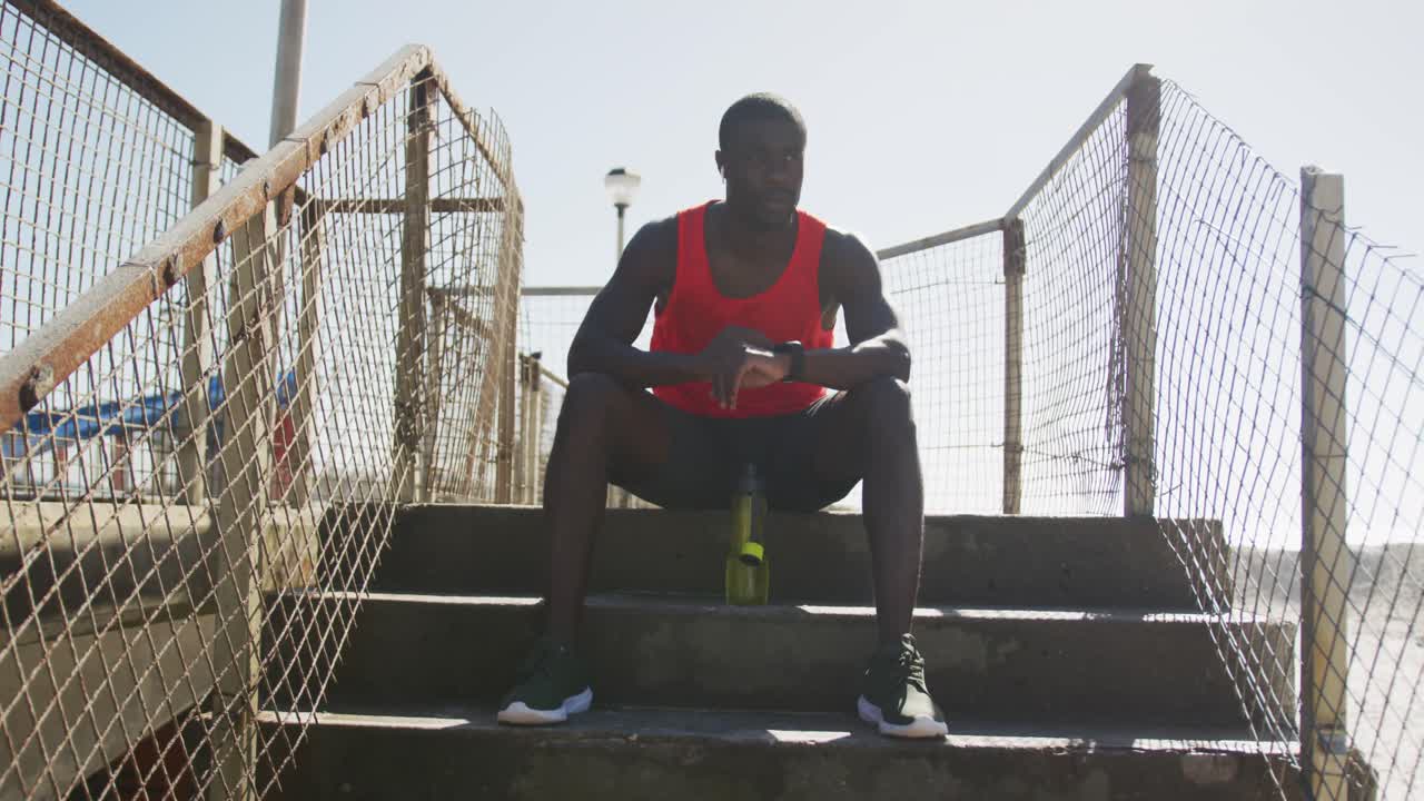African american man sitting, checking his smartwatch, taking break in exercise outdoors