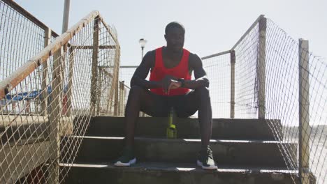 African-american-man-sitting,-checking-his-smartwatch,-taking-break-in-exercise-outdoors