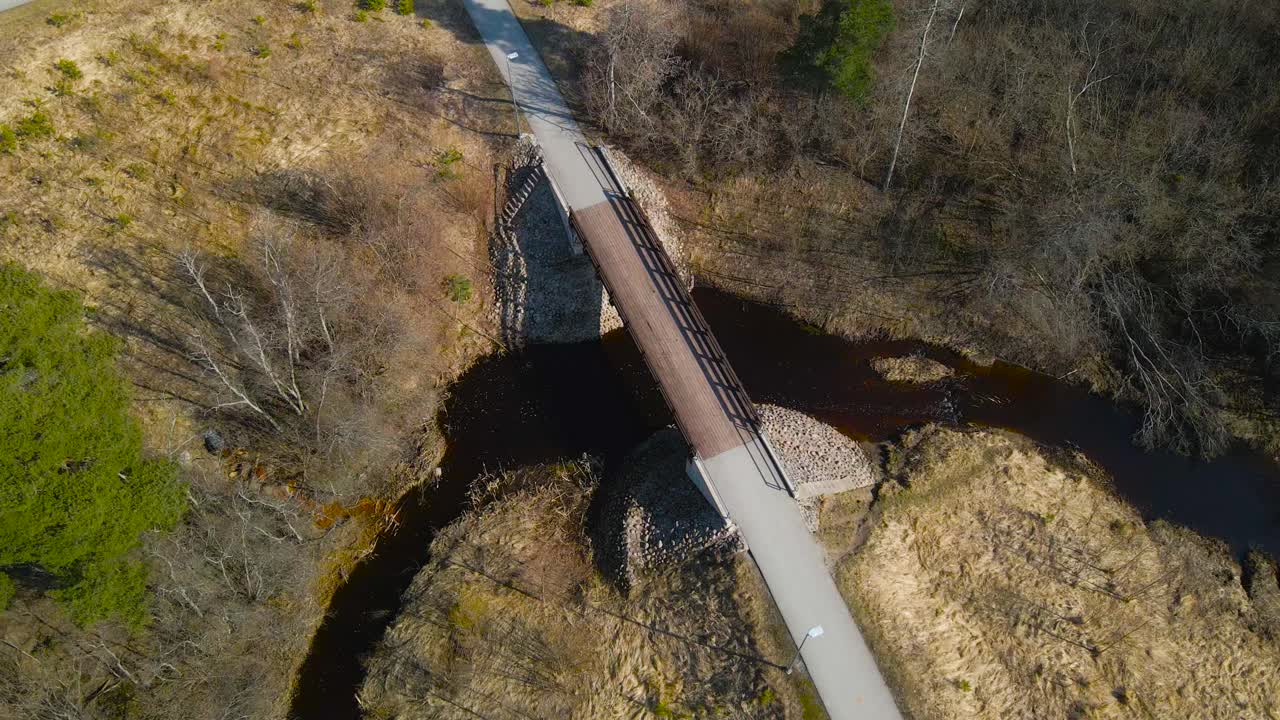 Gorgeous aerial drone footage view of a rural wooden brown bridge in Laagri going over Pääsküla dark colored river during a sunny day in Estonia. The surronding riverbanks are covered in brown grass.