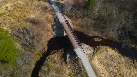 gorgeous aerial drone footage view of a rural wooden brown bridge in laagri going over pääsküla dark colored river during a sunny day in estonia. the surronding riverbanks are covered in brown grass.