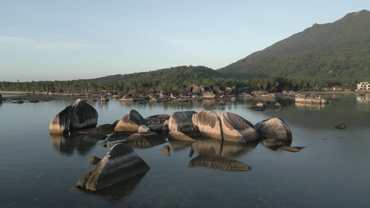 Elokuvamainen matalan lennon dronekuva liukuu Alif Stone Parkin tyynien vesien yllä auringonnousun aikaan. Suuret graniittilohkareet ovat tyynessä meressä rehevää vuoristomaisemaa vasten Indonesiassa.