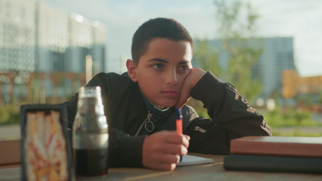 Boy thoughtfully sketching on paper with head resting on hand as adult offers chips and juice on wooden table, outdoor setting with calm mood and subtle focus on study and snacks