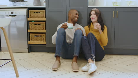 multiracial couple sitting on kitchen floor, laughing and enjoying coffee together, at home