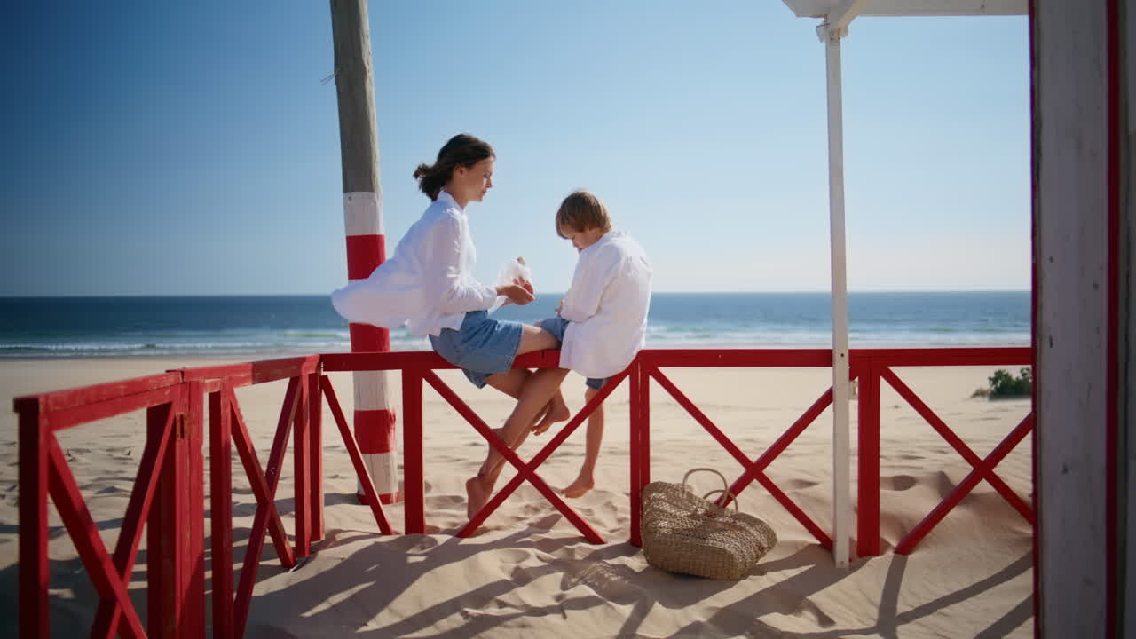 Happy woman talking son sitting beach red fence sunny day. Relaxed mother child