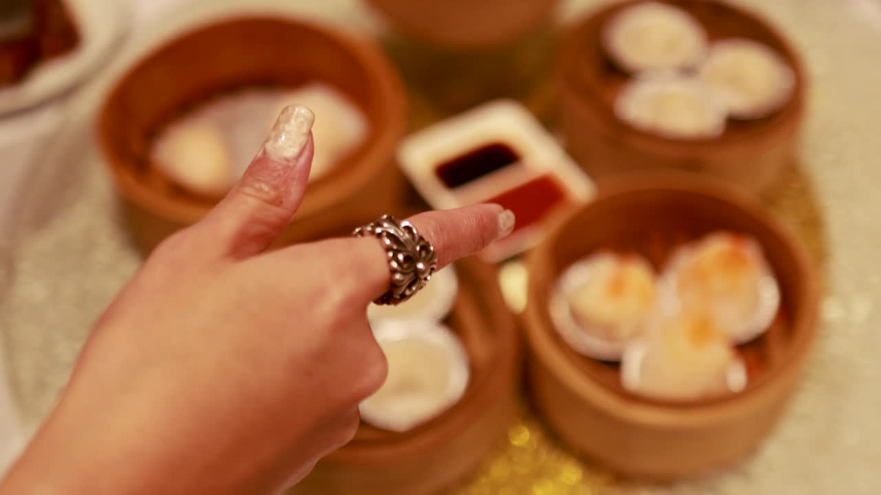Hands choosing dim sum from bamboo steamers on a table with sauces, under warm lighting