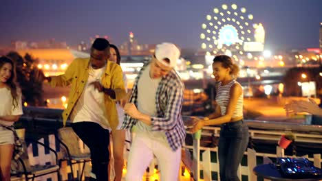 Joyful friends girls and guys are dancing at night party on roof relaxing and laughing when DJ is mixing music using special equipment. Friendship and fun concept.