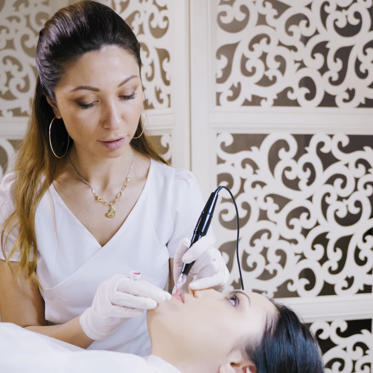 Procedure of permanent make-up for lips of attractive woman in a beauty salon. Pretty young cosmetologist is doing pink tattoo on lips by special machine.