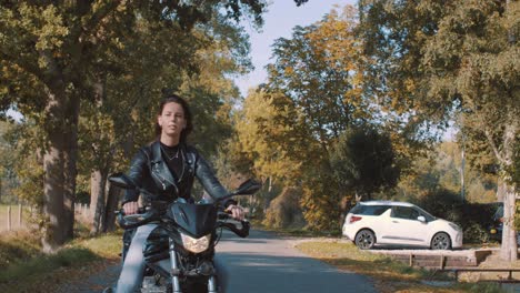 una joven europea muy sonriente conduciendo una moto con una chaqueta de cuero en el bosque con hojas de otoño doradas vibrantes y coloridas en un día soleado