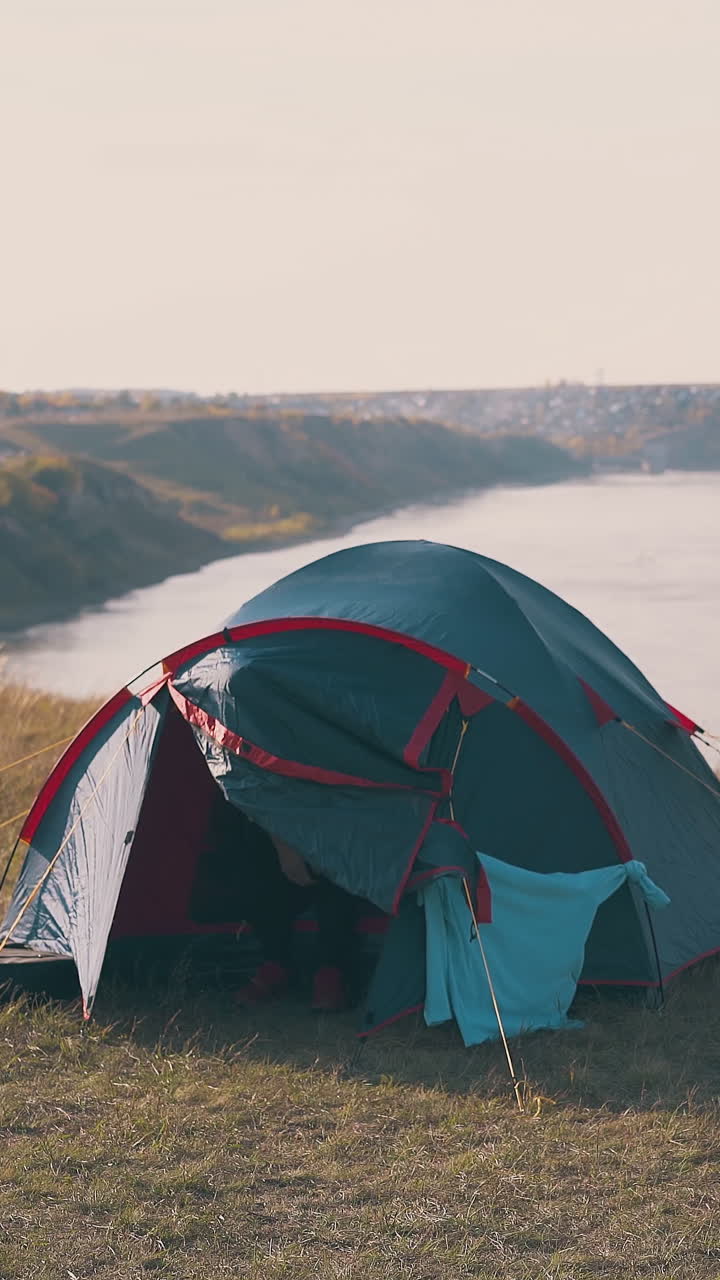 young tipsy man goes out of tent and group of girls near comfortable camp against river in autumn morning