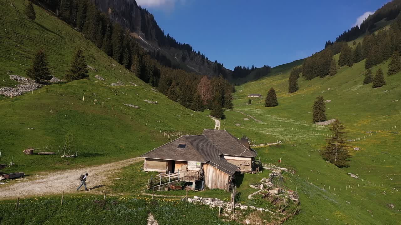 aéreo, mochilero masculino caminando hacia una cabaña rural junto a las montañas durante el verano