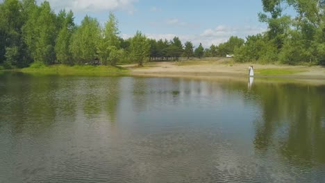 newly-wedded-couple-stands-on-bank-of-tranquil-lake-aerial