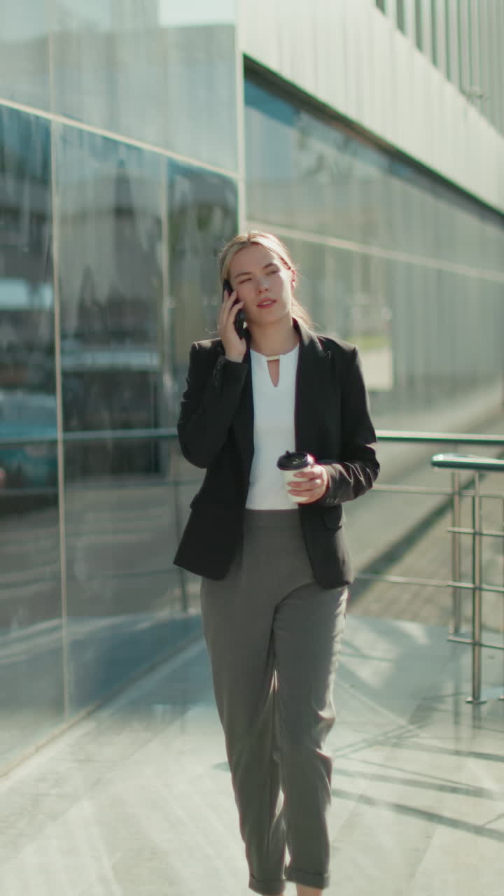 Professional banker walking confidently with coffee in hand on phone call near glass wall and iron railing, reflecting urban scenery and cars, showcasing business lifestyle in modern environment