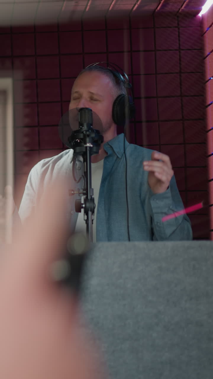 un hombre canta apasionadamente en un estudio de grabación, con auriculares negros y una camisa de manga larga azul. sus ojos están cerrados mientras actúa, gestos con las manos, capturados a través de un espejo. fondo rojo