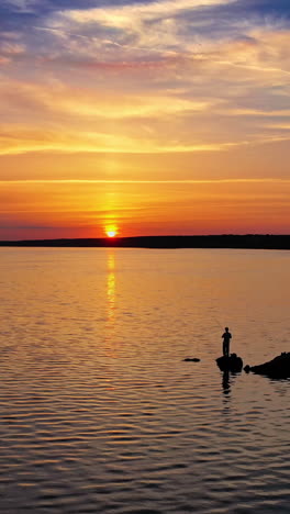 silhouette of a fisherman with fishing rod standing on stone near the river at sunset. evening background over the water and people having great time at the river. aerial view. vertical video