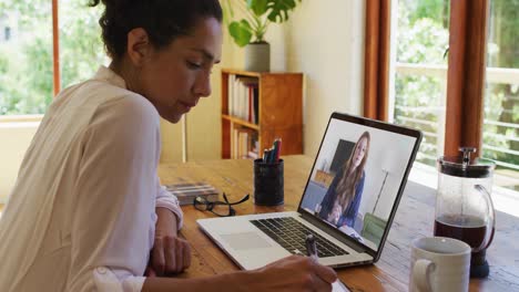 mujer afroamericana tomando notas mientras tiene una videollamada en una computadora portátil en casa