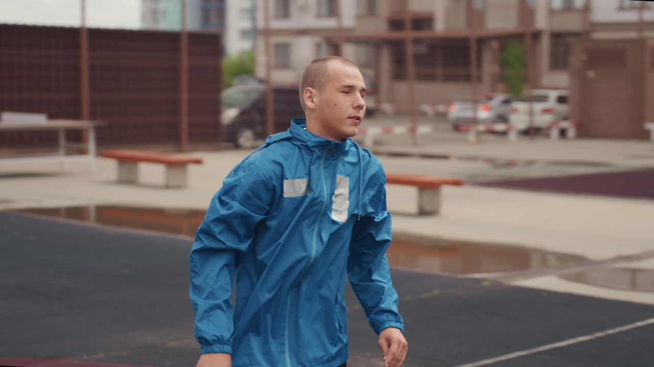 young man in blue rain jacket on wet football court, arms crossed, goalpost in background, puddles on pitch, cloudy sky, coach presence, determined expression, gritty urban training atmosphere, frames