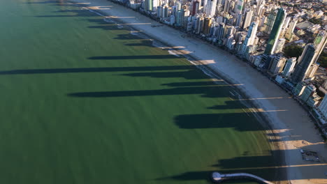 Slow aerial glide capturing long shadows cast by skyscrapers along the coastline of Balneario Camboriu, Santa Catarina, Brazil, creating a striking pattern on the beach and ocean at sunset