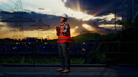 full body side view of asian male engineer with safety helmet smiling and clapping his hands while standing near high voltage tower