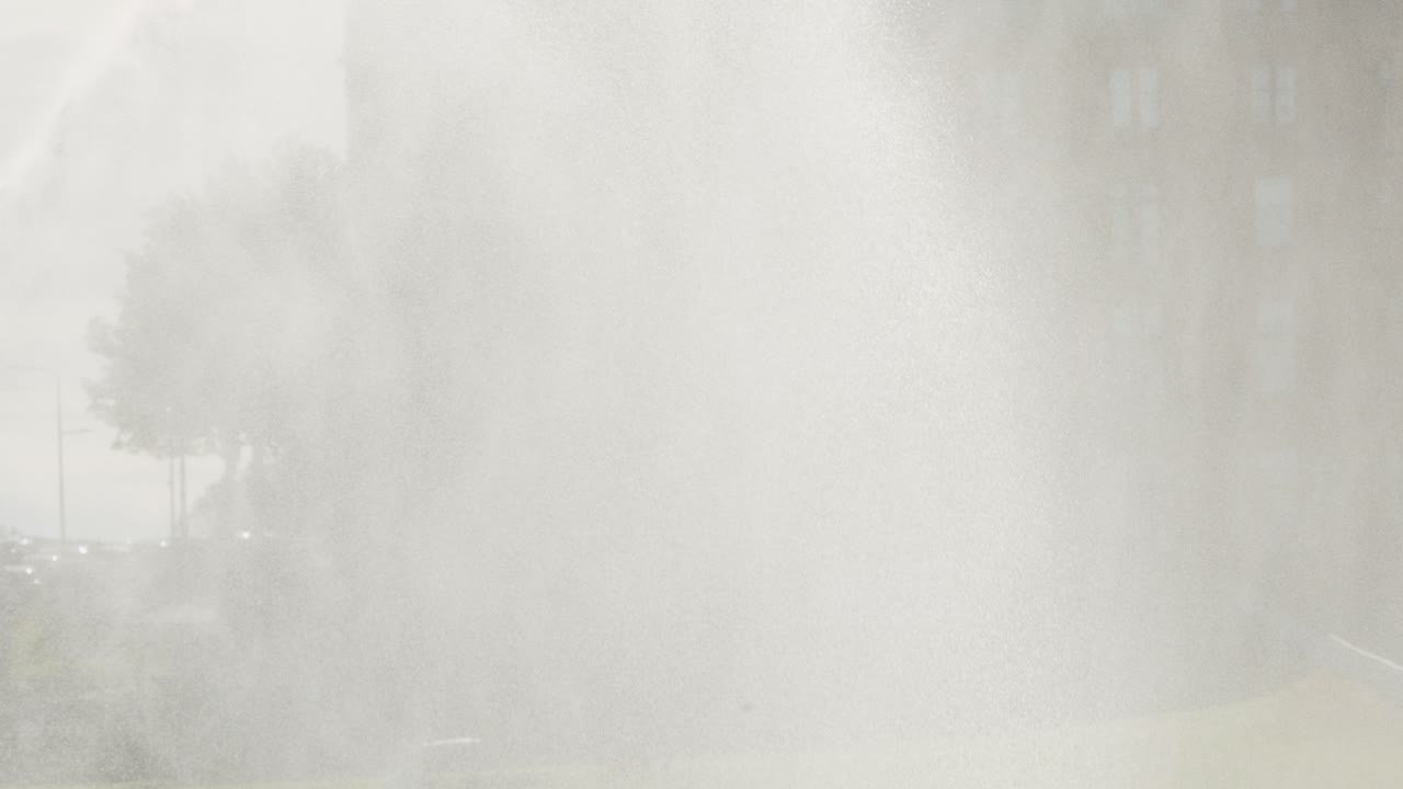 Dense water spray from a golf course sprinkler envelops a flag and surrounding greenery, with soft daylight and misty atmosphere, captured in a static wide shot