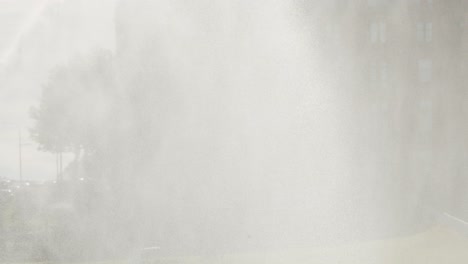 Dense water spray from a golf course sprinkler envelops a flag and surrounding greenery, with soft daylight and misty atmosphere, captured in a static wide shot