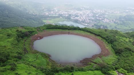 Cinematic-aerial-view-of-the-sacred-lake-at-the-Brahmagiri-hill-in-the-Western-Ghats-of-Maharashtra-during-monsoon-with-the-view-of-Trimbak-town-in-the-background,-Nashik,-India