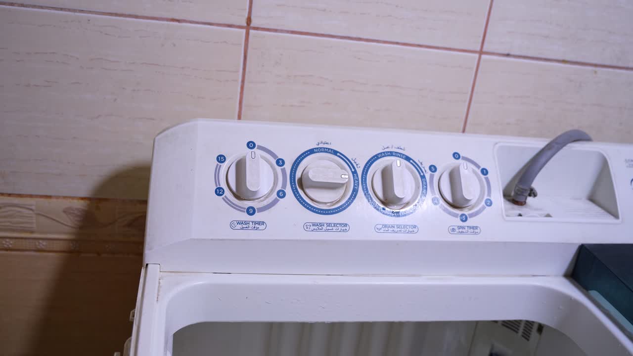 Close-up of a man's hand adjusting the selector dial on a top-loading washing machine control panel. Features labels in both English and Arabic for wash settings