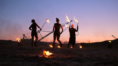 a group of men and woman fire show at night on the sand against the background of fire and tower cranes.