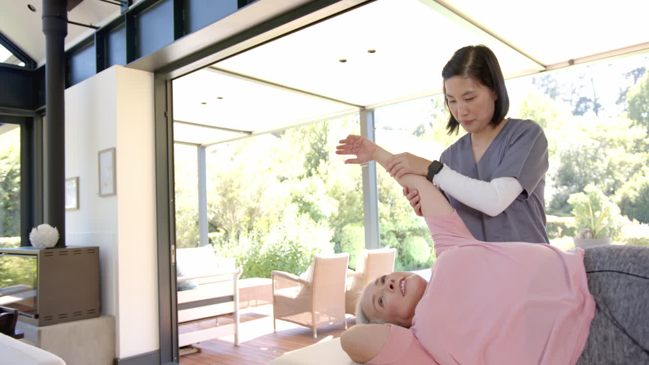 Assisting senior woman with arm exercises, physiotherapist working at home