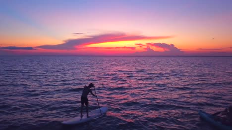 Eine-Dramatische-Drohnenaufnahme-Eines-Kajakfahrers-Und-Paddelboarders,-Der-Langsam-In-Einen-Farbenfrohen-Sonnenuntergang-Hineindriftet