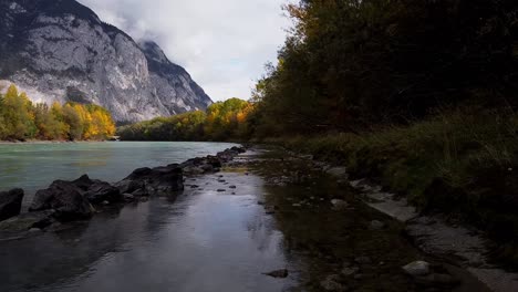 Flying-along-side-of-river-with-trees-growing-around,-mountains-in-the-distance-on-a-sunny-day