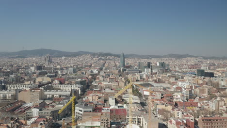 Fly-over-construction-site,-group-of-tall-tower-cranes.-Cityscape-with-hilly-landscape-in-background.-Barcelona,-Spain