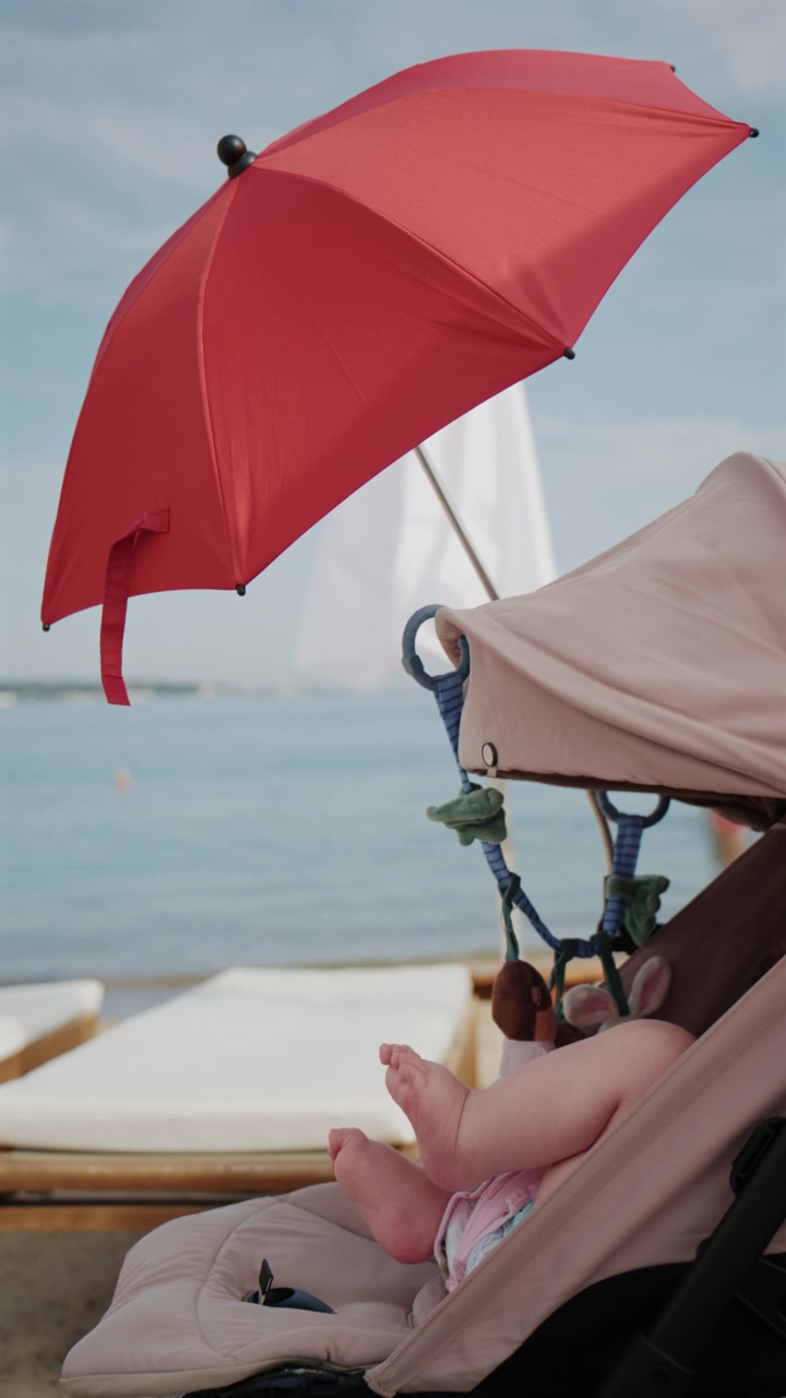 Bambino seduto nel passeggino sotto un ombrellone da spiaggia rosso brillante in riva al mare.