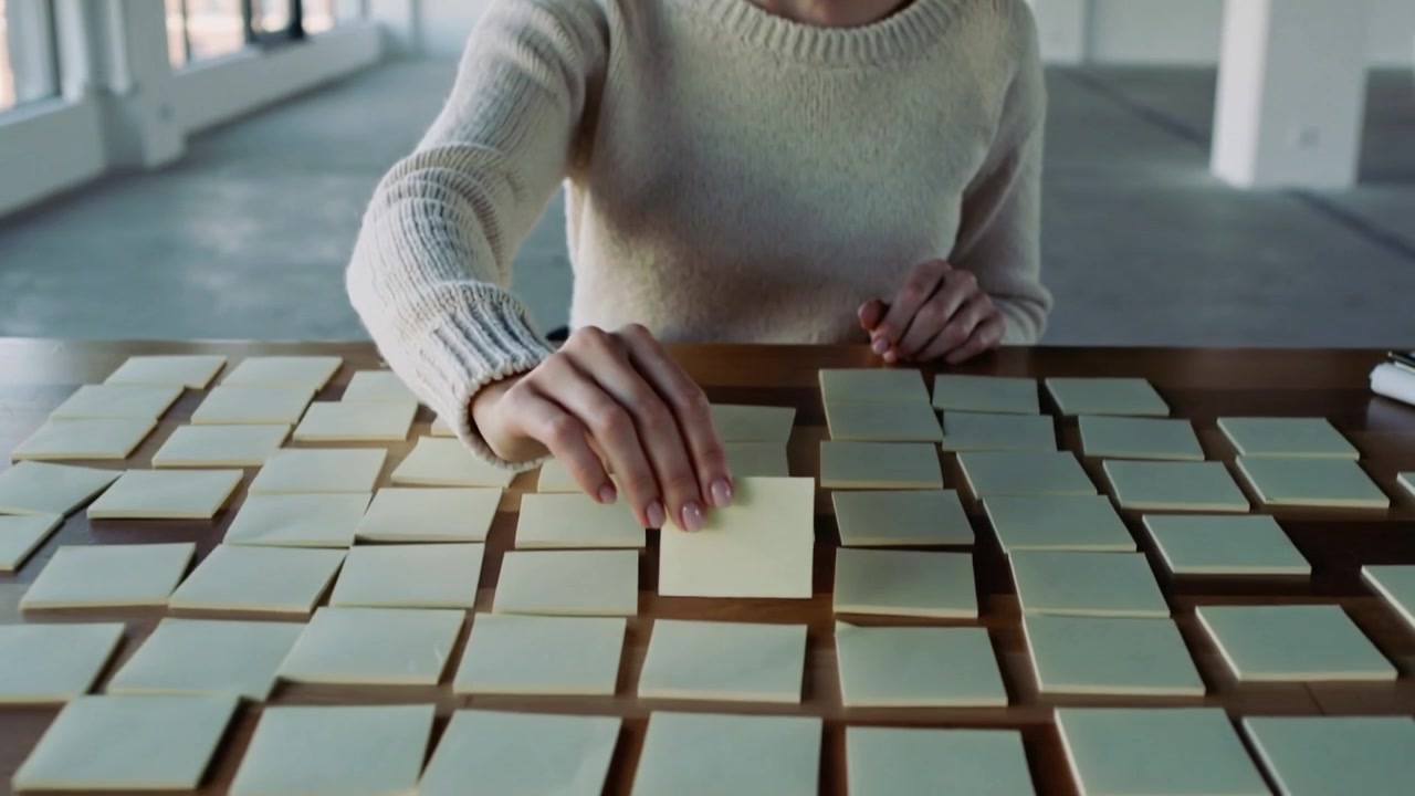 Scanning sticky note grid, woman in light sweater moving notes at loft table organizing plan