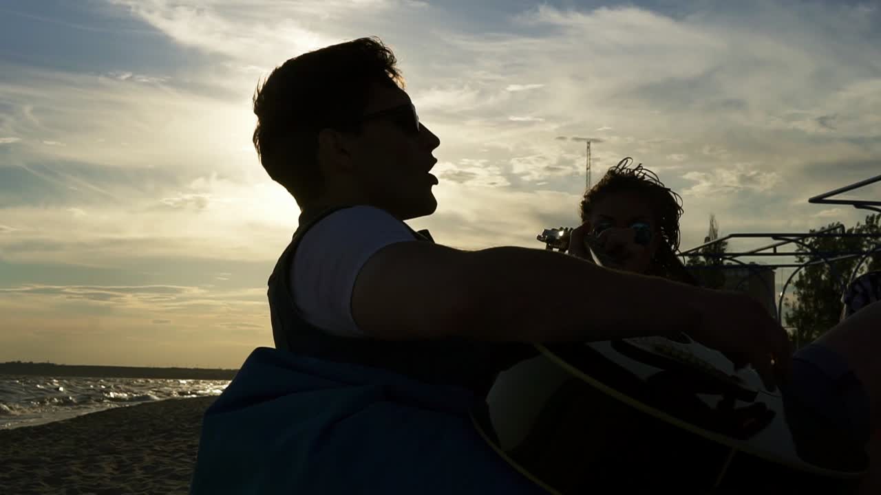 해가 지는 여름 저녁에 해변에 앉아서 편안한 의자에 앉아서 기타를 연주하는 젊은 남자. 슬로우모션  ⁇