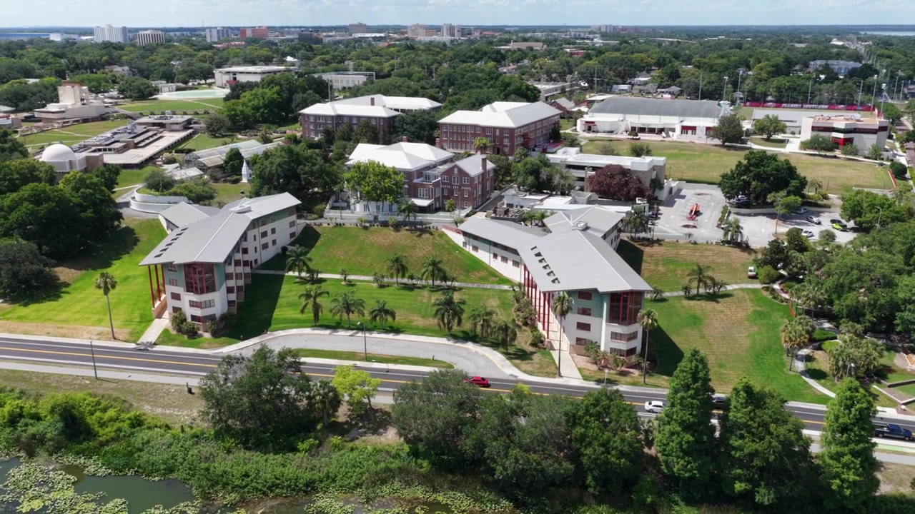 Campus del Florida Southern College che mostra l'architettura di Frank Lloyd Wright a Lakeland, Florida, con passerelle coperte che collegano gli edifici tra palme vicino a una strada lungo il lago. Ripresa aerea con drone in orbita.