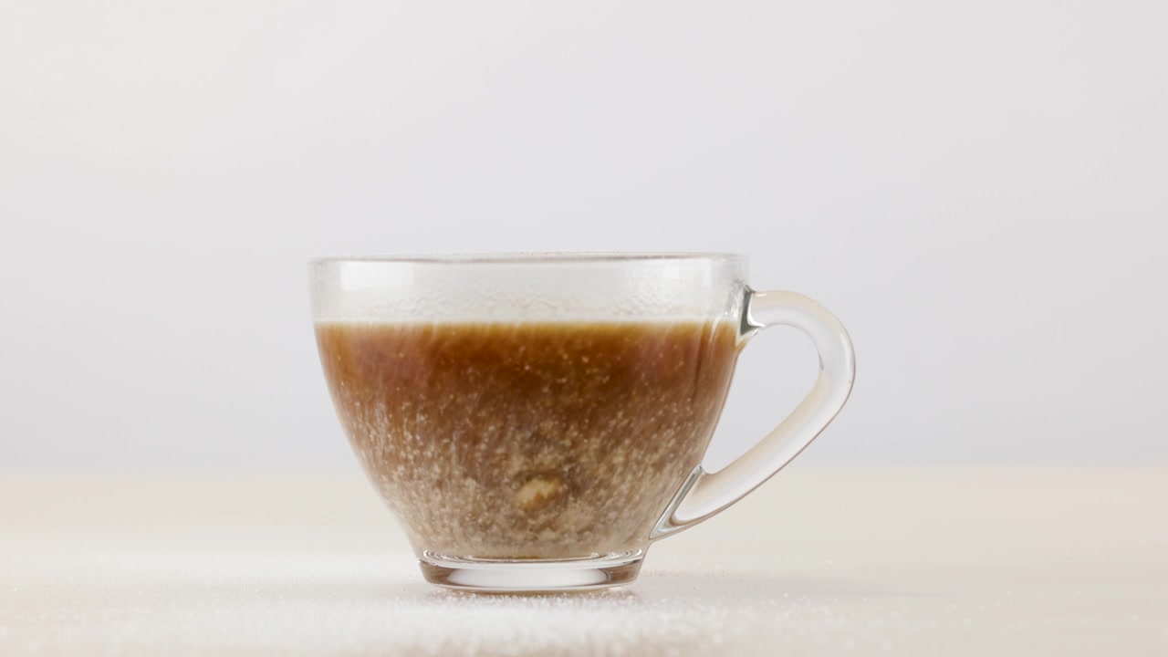 Zucchero semolato cade in una tazza di vetro di caffè espresso su uno sfondo bianco pulito.