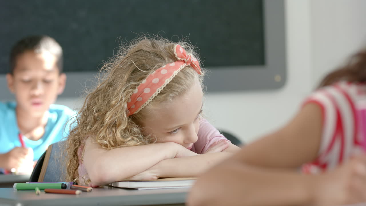 In school, girl resting head on desk while classmates studying in classroom