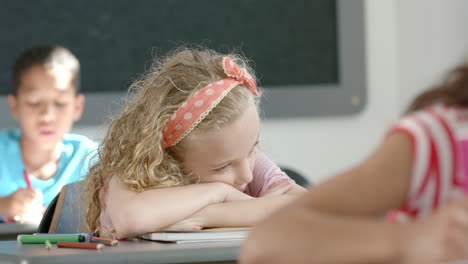 in school, girl resting head on desk while classmates studying in classroom