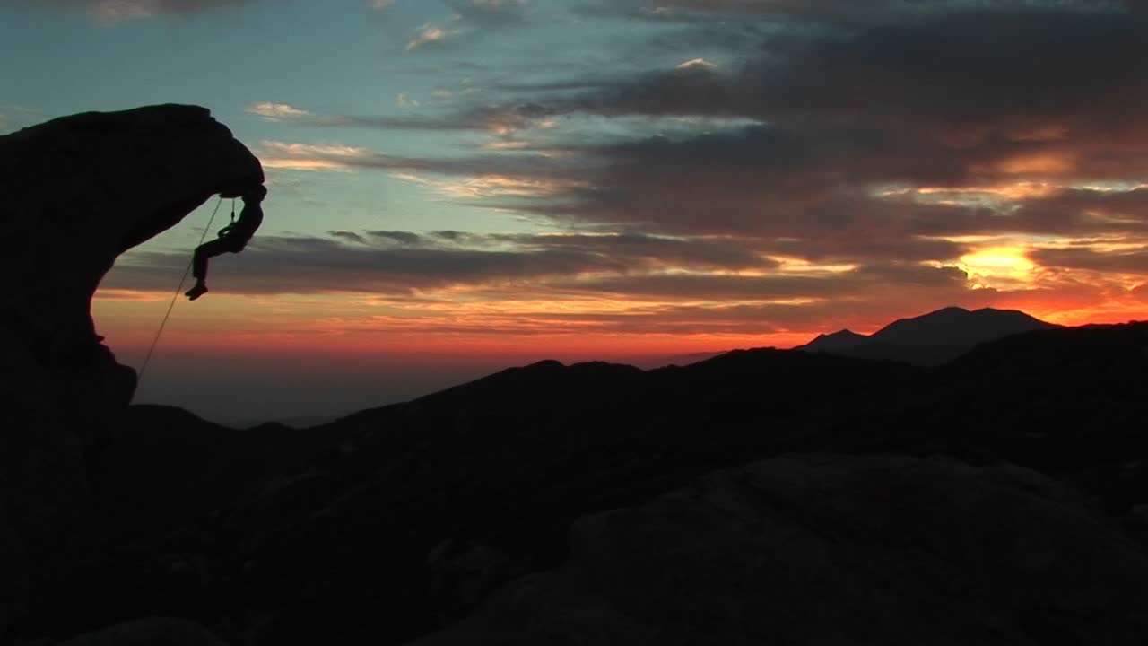 Mezza figura di uno scalatore in silhouette contro il sole al tramonto, appeso a uno strapiombo roccioso.