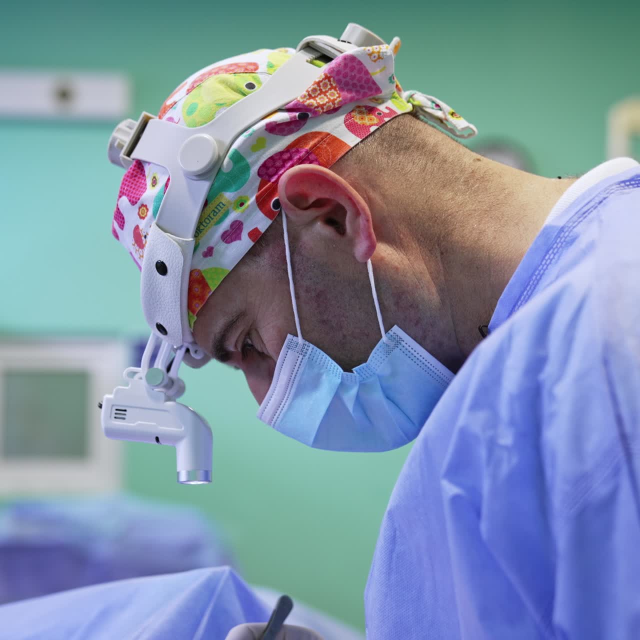 Profesional de la cirugía con máscara, faro y gorro colorido. Vista lateral. Retrato de un médico centrado en la intervención.