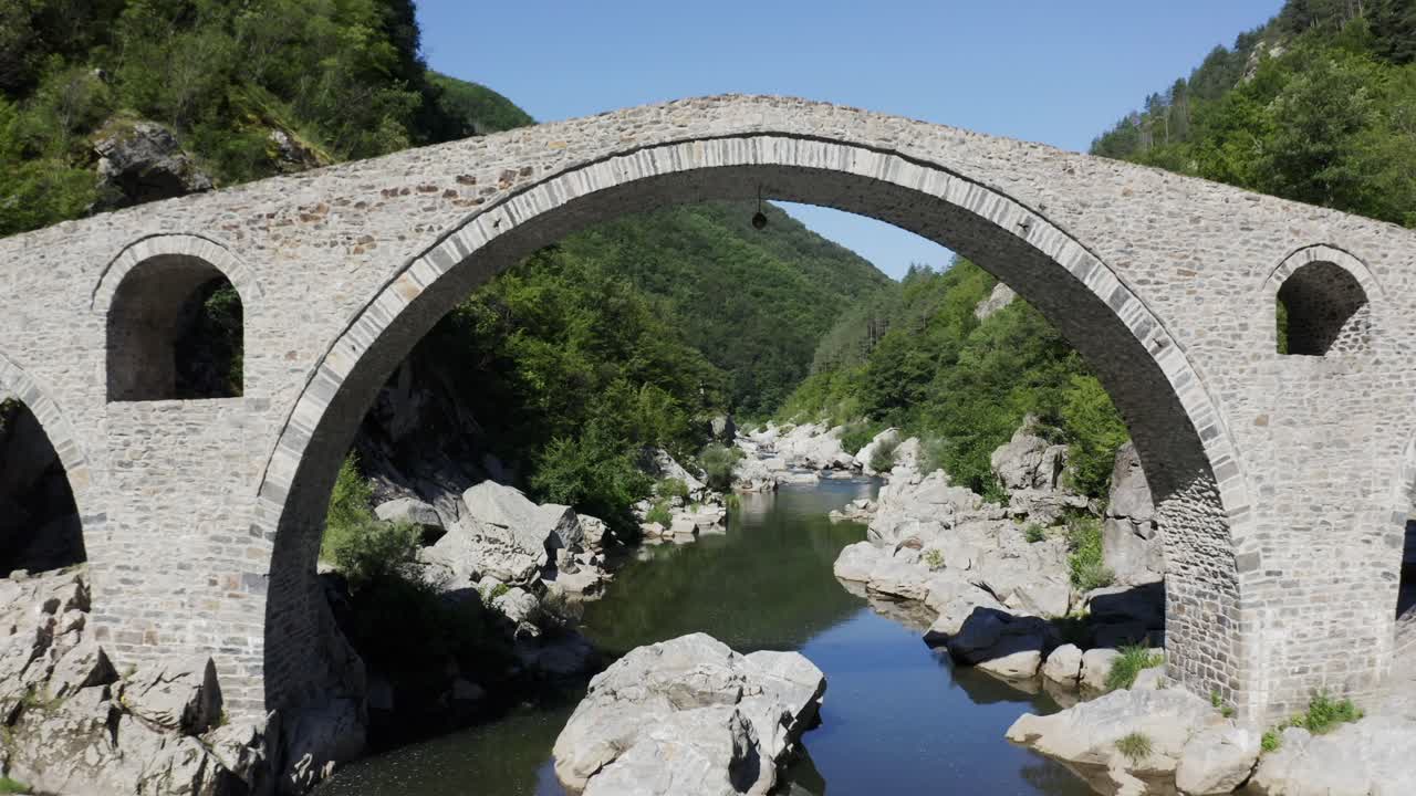 Retreating drone shot over the Arda River and going through the main arch of the Devil's Bridge and revealing the beautiful landscape at the foot of the Rhodope Mountains in Bulgaria