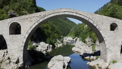 Retreating drone shot over the Arda River and going through the main arch of the Devil&#039;s Bridge and revealing the beautiful landscape at the foot of the Rhodope Mountains in Bulgaria