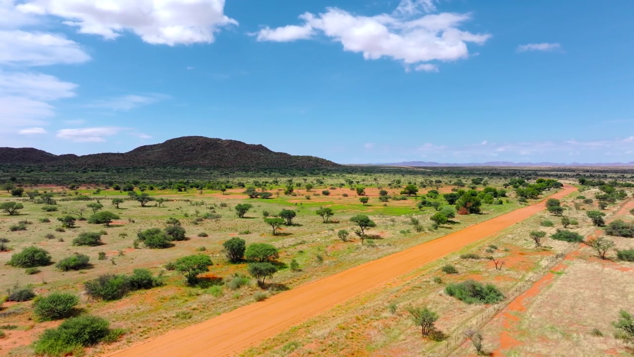 Vista drone di una strada di sabbia rossa che attraversa la boscaglia verde del Kalahari punteggiata da alberi di acacia verso una bassa collina. Sabbia rossa infinita ed erba dorata raggiungono l'orizzonte