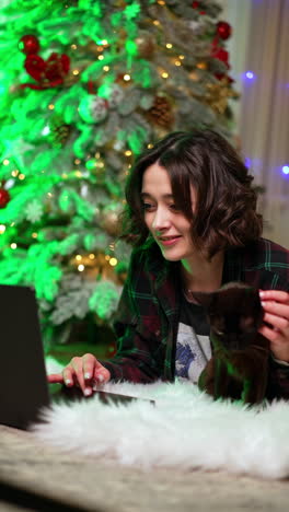 smiling caucasian woman lying on the fluffy plaid holding a black cat and working on laptop. beautiful christmas tree at backdrop. vertical video.