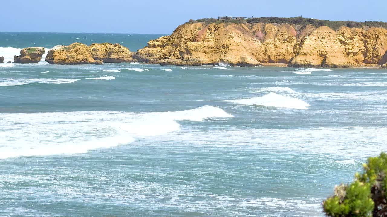 Ocean waves crash against rocky cliffs under clear skies in Torquay, Victoria. Bright daylight enhances the vibrant coastal scenery